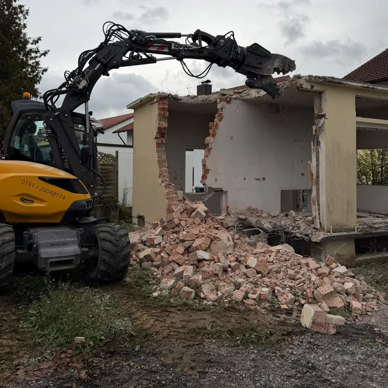 Mobilbagger mit Abbruchzange reißt die Außenwand eines Anbaus ein, davor ein Haufen Ziegelschutt